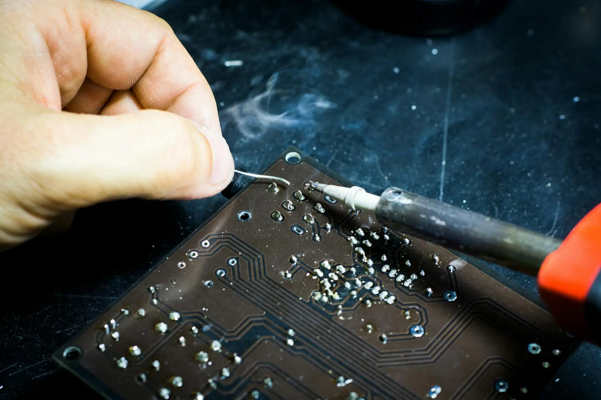 Kaviapps-wordpress-Plugin-Customization-main-banner Close-up of a hand soldering a circuit board on a dark surface. Smoke rises from the heated area as a soldering iron and solder wire are used to connect components, highlighting the intricate electronic work.