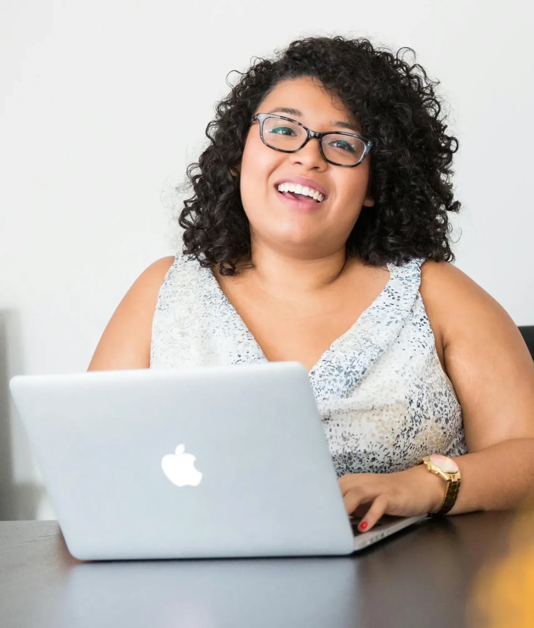 Happy client sharing a positive testimonial A women with curly hair and glasses is smiling while sitting at a desk using a laptop. The laptop is silver with an apple logo. They are wearing a patterned sleeveless top.